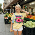 May include: A light yellow t-shirt with the text "SHE RUNS this SUMMER" and a graphic of a flip phone, sunglasses, and a pink hibiscus flower. The person is wearing white shorts and a blue purse.