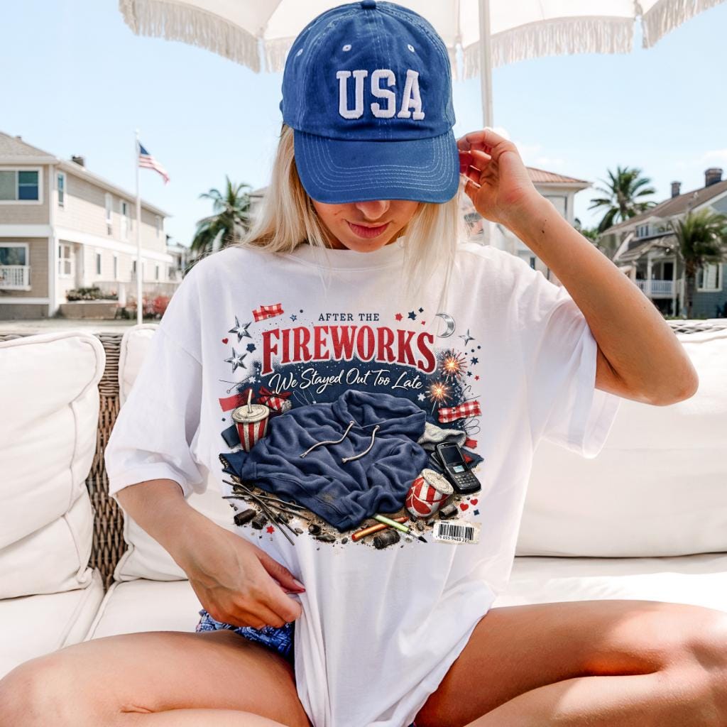May include: White t-shirt with the text "AFTER THE FIREWORKS We Stayed Out Too Late" and a graphic of fireworks, a blue hoodie, and other items. A blue baseball cap with "USA" is worn.